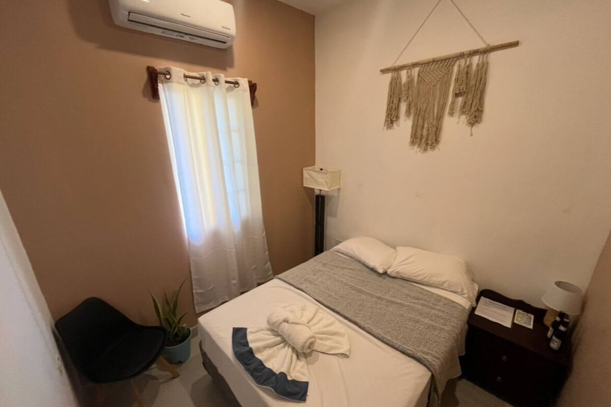 A bedroom with a bed, a bedside table, a lamp, a plant, and a wall-mounted air conditioner.