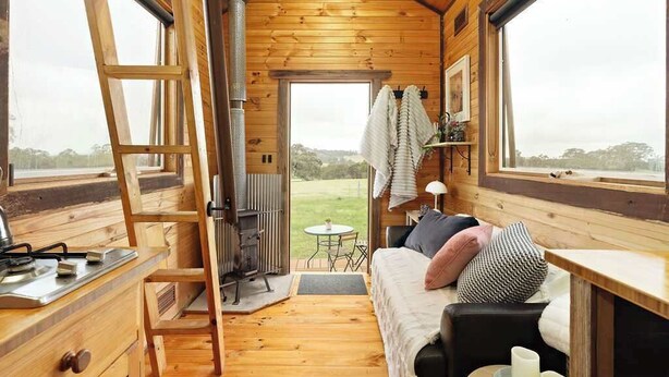 Interior acogedor de una cabaña con cama en altillo, chimenea, comedor y ventanas amplias que ofrecen vistas al exterior.