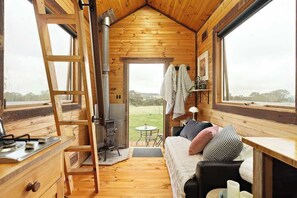 A cozy cabin interior with a loft bed, a fireplace, a dining area, and large windows offering a view of the outdoors.