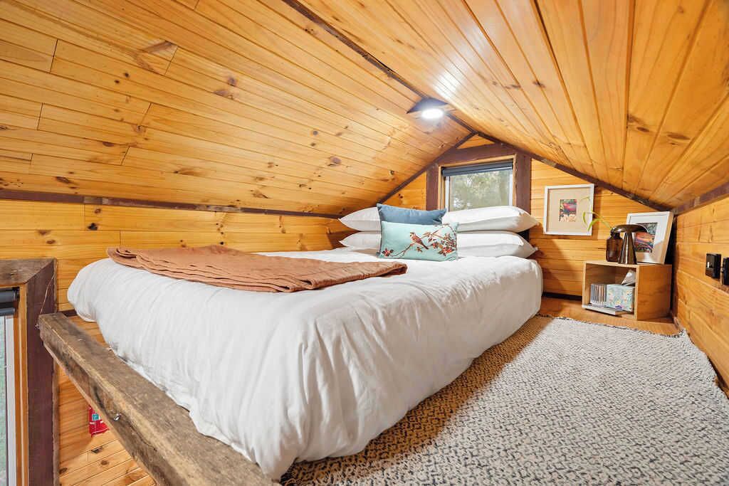 Een gezellige slaapkamer onder de dakpannen, met een houten plafond, een bed met witte beddengoed, een bruine plaid en een klein raam.
