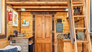 Un interior acogedor de cabaña con puerta de madera, una pequeña kitchenette, cama y escalera.