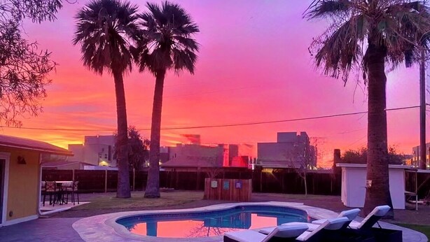 Un jardin avec une piscine, des chaises longues, des palmiers et une vue sur la ville au coucher du soleil.