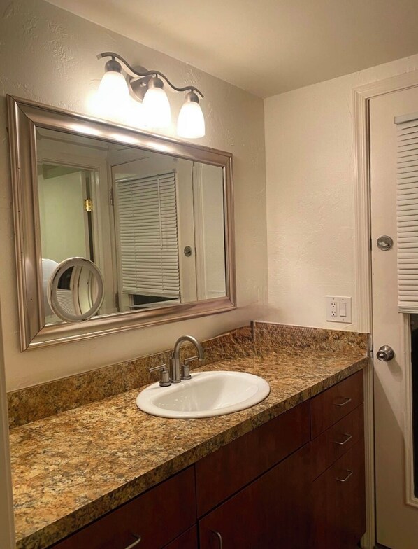 Une salle de bain dotée d’un comptoir en granit, d’un lavabo blanc, d’un miroir avec éclairage intégré et d’une porte avec des stores.