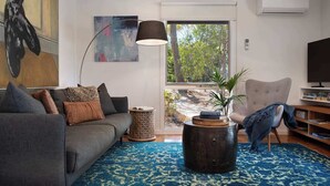 A living room with a grey sofa, a round coffee table, a lamp, a bookshelf, and a view of trees through a glass door. - Kingfisher Loft – 2BR Spa Bath Retreat near Daylesford & Hepburn Springs (Daylesford)