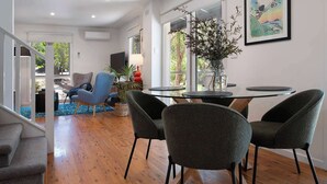 A modern dining area with a round glass table, four grey upholstered chairs, and a large window with a view of greenery. - Kingfisher Loft – 2BR Spa Bath Retreat near Daylesford & Hepburn Springs (Daylesford)