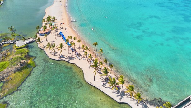 Luchtbeeld van een strand met helder turquoise water, een zandrijk strand langs palmbomen en enkele boten in de verte.