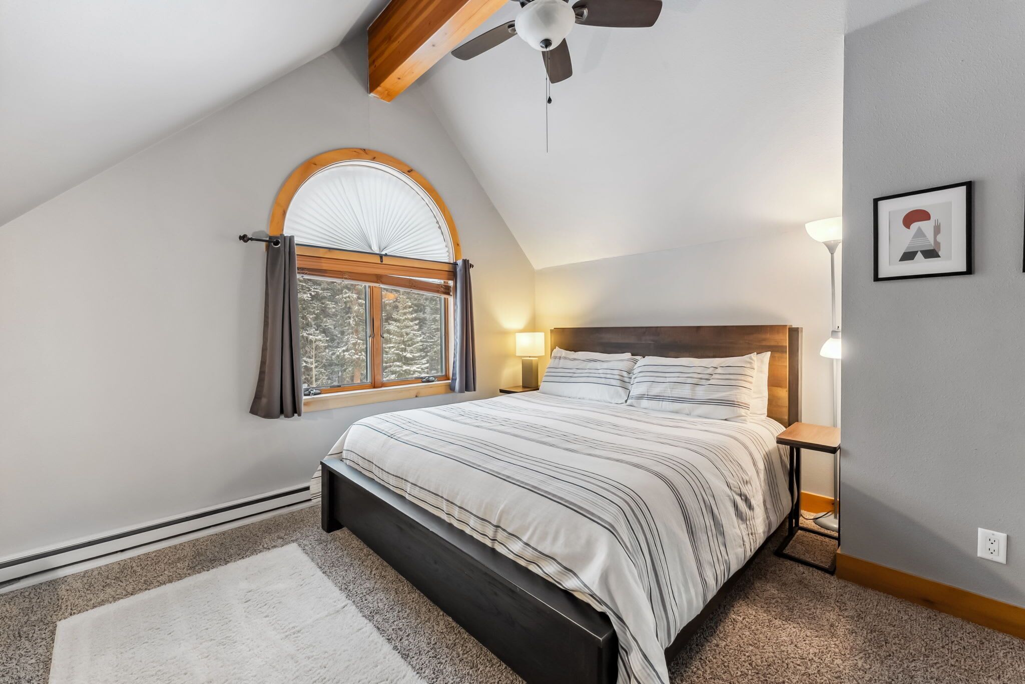 Une chambre avec un plafond en pente, un lit doté d’une tête de lit en bois, une fenêtre avec des rideaux, une lampe de chevet et un tableau encadré accroché au mur.