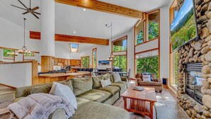 Ein geräumiges Wohnzimmer mit einer großen Couch, einem Couchtisch und einem Kamin. Es gibt eine Küchenzeile mit Holzausstattung und einen Esstisch. Der Raum verfügt über große Fenster und eine Steinwand.