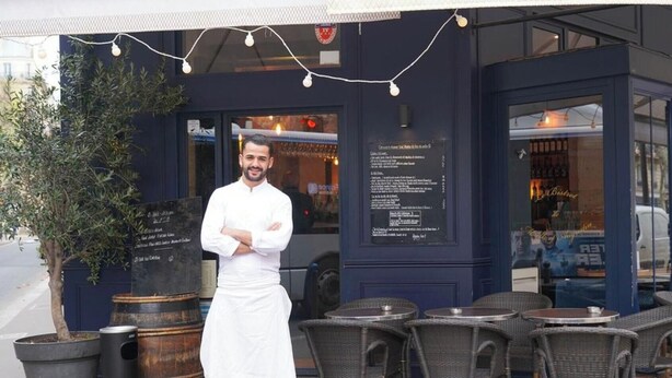 Un chef de pie frente a un restaurante con terraza, una pizarra de menú y una planta en maceta.