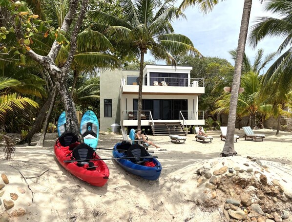 Un moderno edificio de dos pisos con balcón, kayaks y sillones de playa sobre una arena.