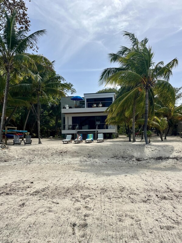 Un edificio moderno frente a la playa, con techo plano, ventanas amplias y un balcón. Varias sillas de descanso y una sombrilla azul están sobre la arena.