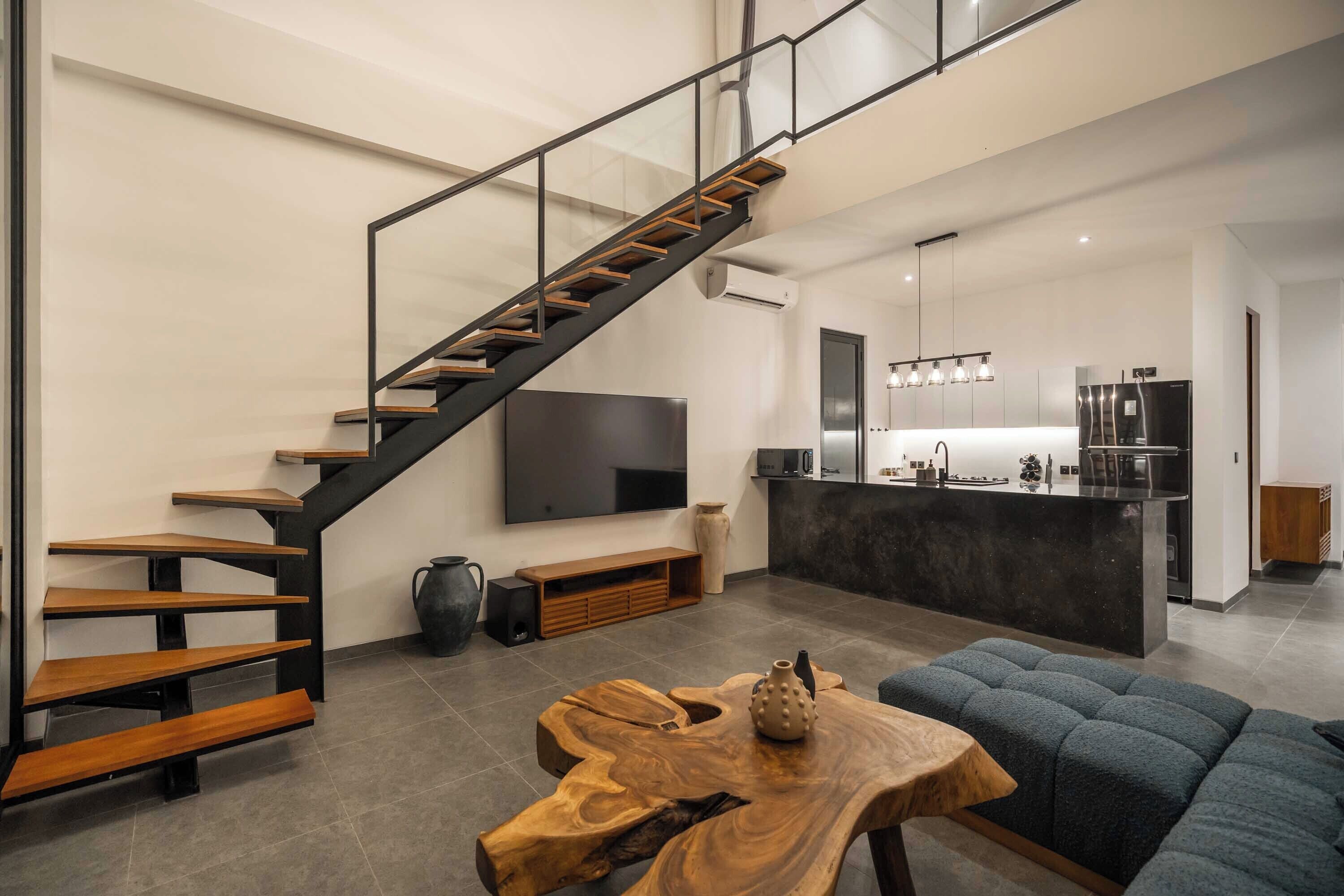 Uma sala de estar moderna com escada de madeira, uma televisão grande e uma área de cozinha.