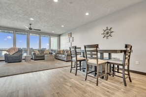 Un salon spacieux doté d’une grande fenêtre, d’un ventilateur de plafond et d’un coin repas avec des chaises et une table en bois.
