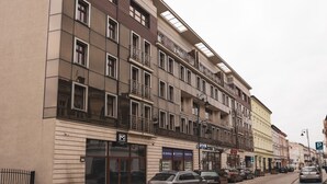 Een meerlaags gebouw met balkons, een inkom op de begane grond en geparkeerde auto's langs de straat.