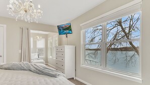A bedroom with a large bed, a dresser, a TV, and a window with a view of trees.