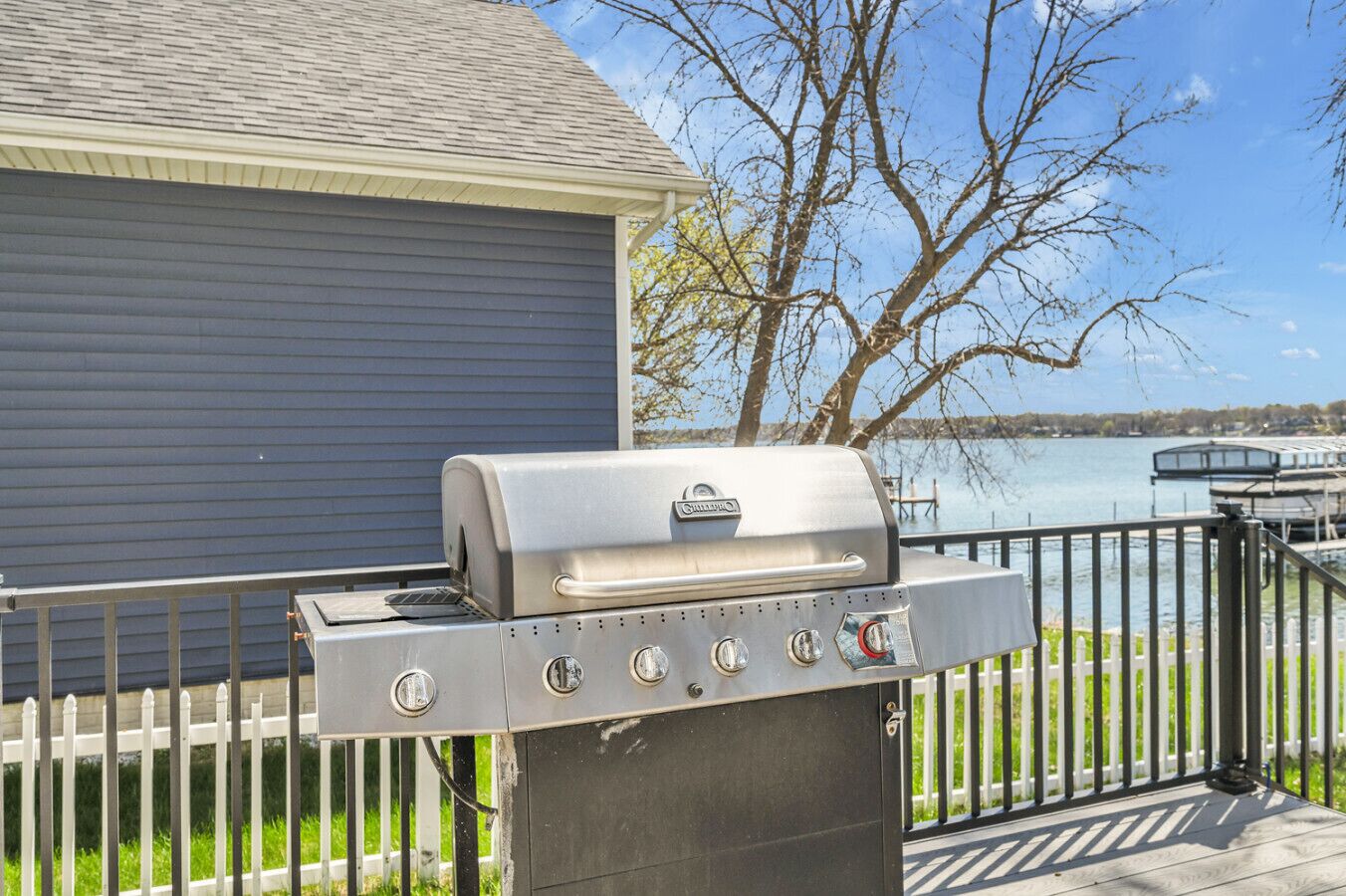 Een roestvrijstalen barbecue met verschillende bedieningsknoppen, op een terras met een reling, nabij een huis en een waterpartij.