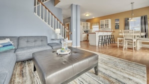A spacious living area with a grey sofa, a leather ottoman, and a kitchen in the background.