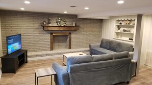 Un salon avec un mur en briques, une cheminée, un canapé gris, un téléviseur posé sur un meuble et des étagères ornées d’objets décoratifs.