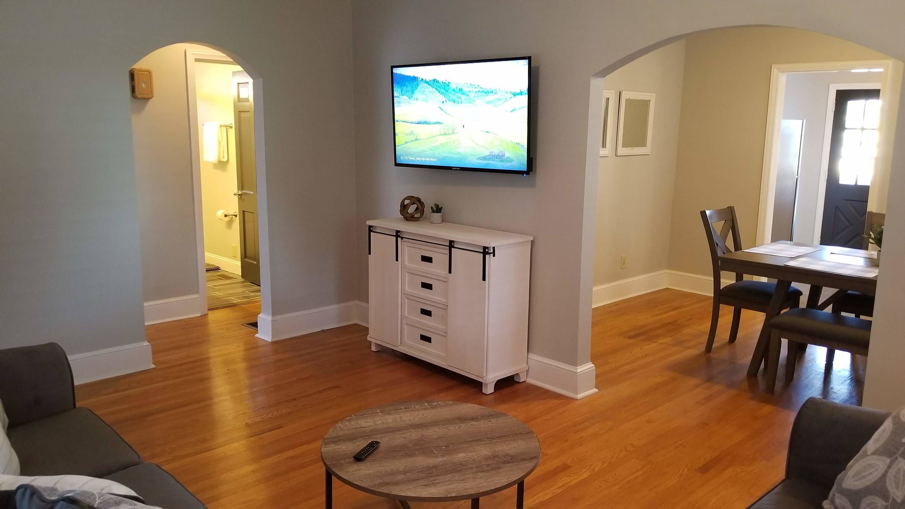 Una sala de estar con piso de madera, un mueble blanco para el entretenimiento, un televisor de pantalla plana montado en la pared, una mesa de centro redonda de madera, y un comedor con mesa y sillas.
