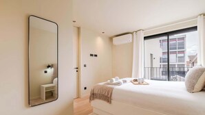 A bedroom with a large bed, a bedside table, a mirror, and a window with a view of buildings.