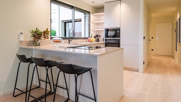 A modern kitchen with a central island, black bar stools, a large window, and built-in appliances.