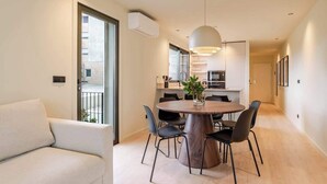 A modern apartment with a dining area featuring a round wooden table and black chairs, a sofa, and a large window with a view of the city.