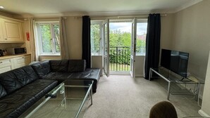 Ein modernes Wohnzimmer mit einem schwarzen Ledersofa, einem Glastisch, einem Flachbildfernseher und einem Balkon mit Blick auf Grünflächen.