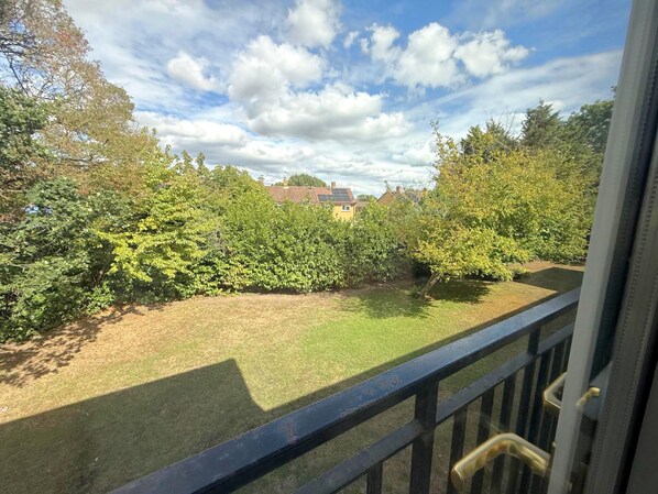 A view from a balcony with a metal railing, a grassy lawn, dense shrubbery, and a house with a solar panel in the background.