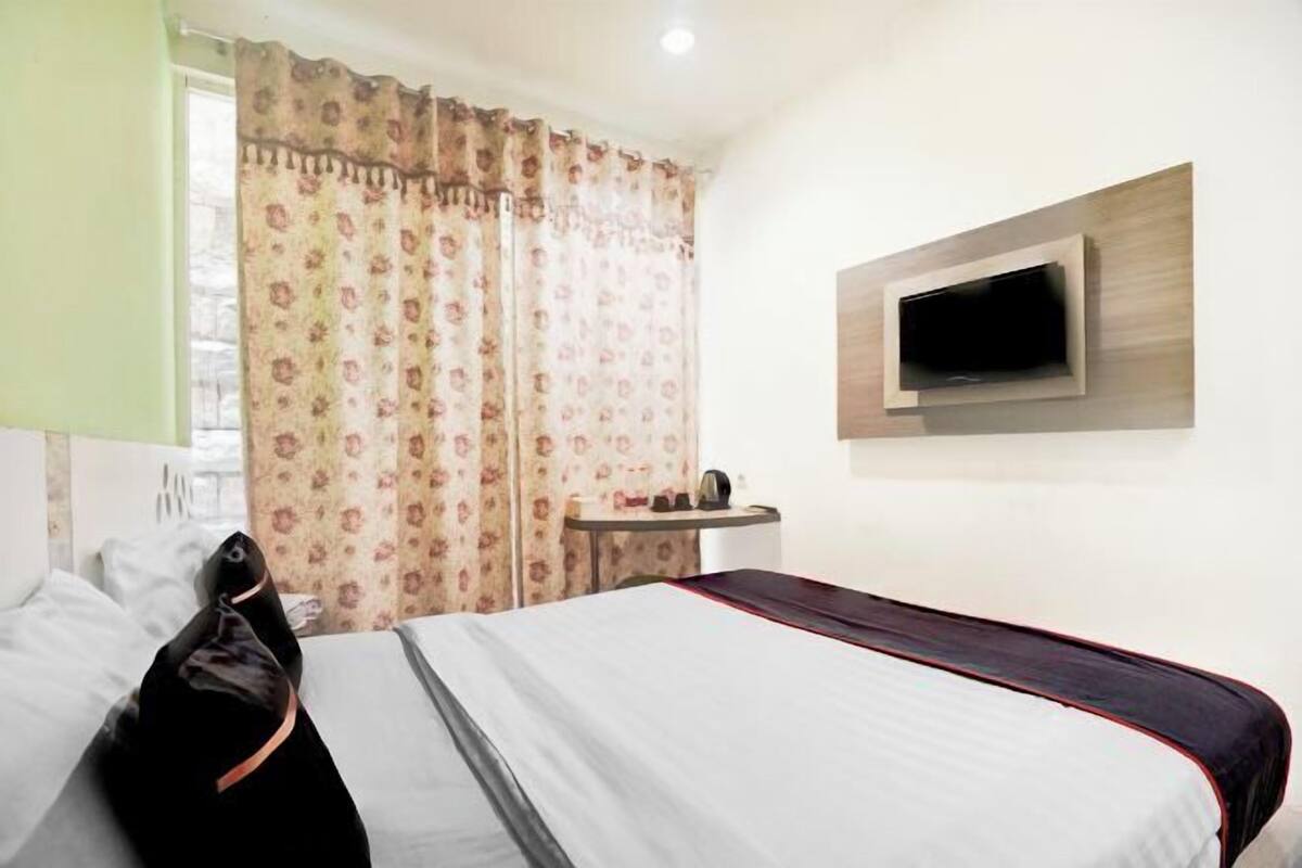 Een hotelkamer met een bed, een aan de muur bevestigde televisie, een klein tafeltje en een raam met gordijnen.