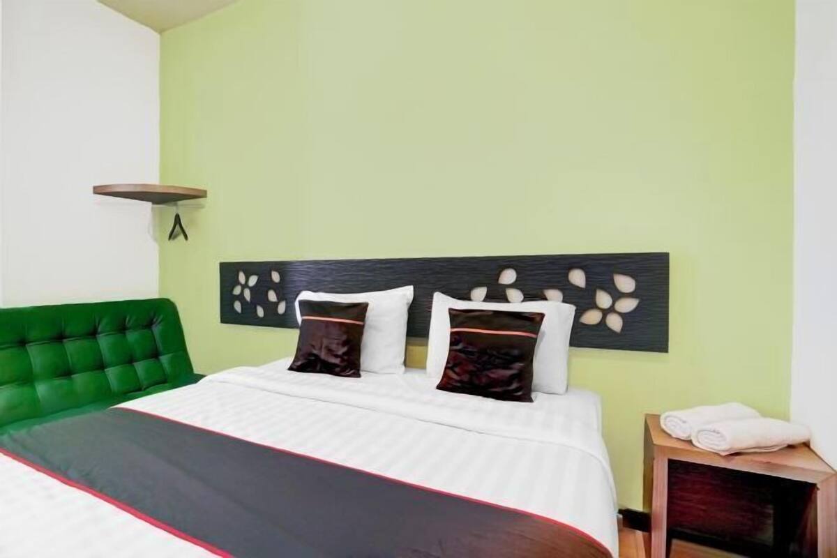Een hotelkamer met een groen gestoffeerd hoofdeinde, een bed met witte lakens en twee decoratieve kussens, een houten nachtkastje met een opgerolde handdoek, en een wandplank.