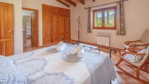 Een slaapkamer met een groot bed, een houten garderobe, een schommelstoel en een raam met uitzicht op groen.