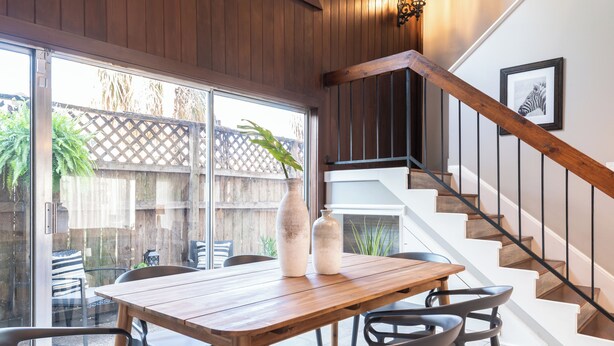 Un comedor con mesa y sillas de madera, una escalera con barandilla de madera y vistas a un patio exterior con plantas a través de un vidrio.