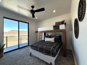 Une chambre avec un lit superposé, un ventilateur de plafond, une grande fenêtre donnant sur un paysage désertique et une plante en pot.