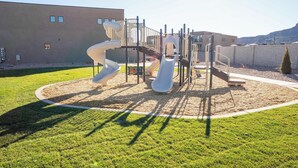 Ein Spielplatz mit Rutschen und Klettergerüsten, umgeben von einer Rasenfläche und einem Gebäude im Hintergrund.