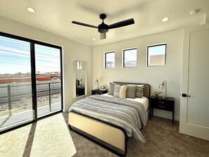 Ein Schlafzimmer mit Bett, Nachttisch, Kommode, Deckenventilator und einem großen Fenster mit Blick auf ein Gebäude und einen Zaun.