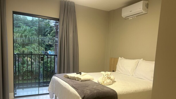 A hotel room with a bed, a window with a view of greenery, and an air conditioning unit.
