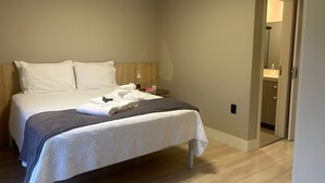 A neatly made bed with white linens and a gray blanket, a bedside table with a book and a remote control, and a wall-mounted air conditioning unit.