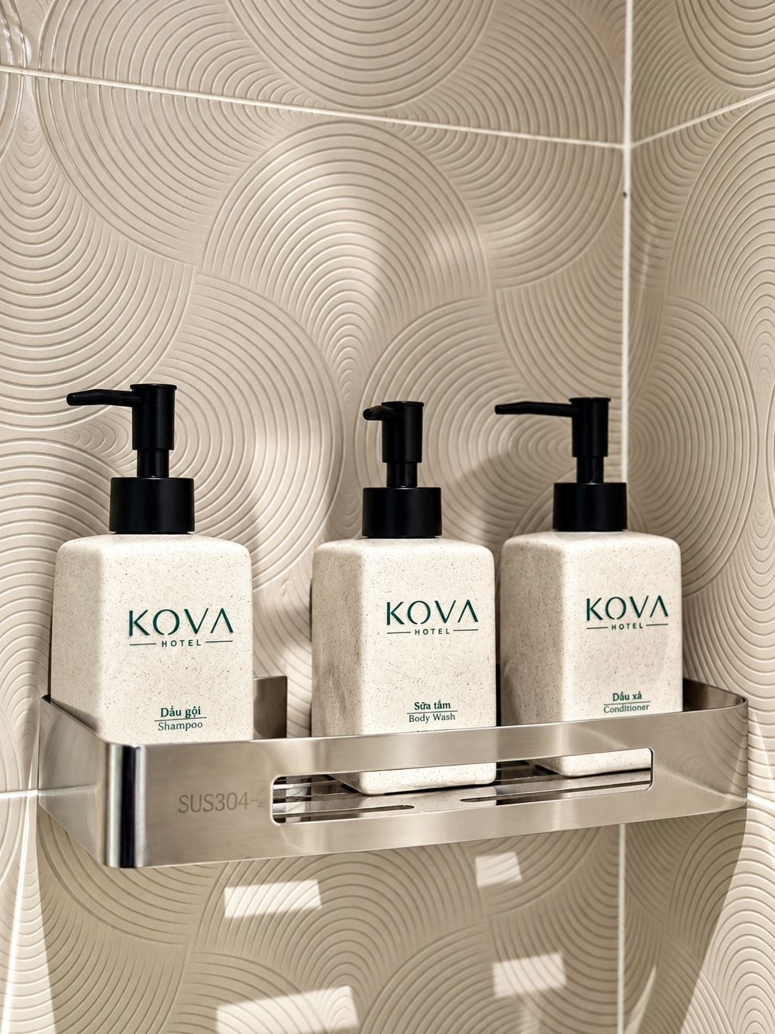 A bathroom with a textured wall and a chrome shelf holding three KOVA Hotel products: shampoo, body wash, and conditioner.