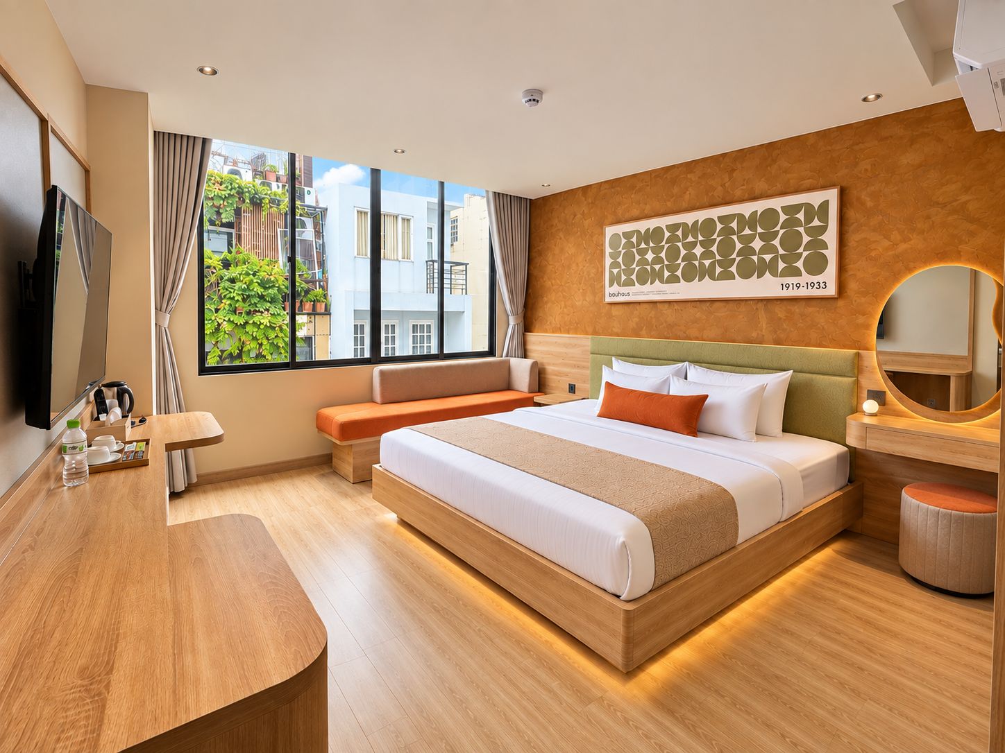 Ein Hotelzimmer mit Bett, Schreibtisch, Stuhl, einem Fenster mit Blick auf Gebäude und einem an der Wand befestigten Kunstwerk.