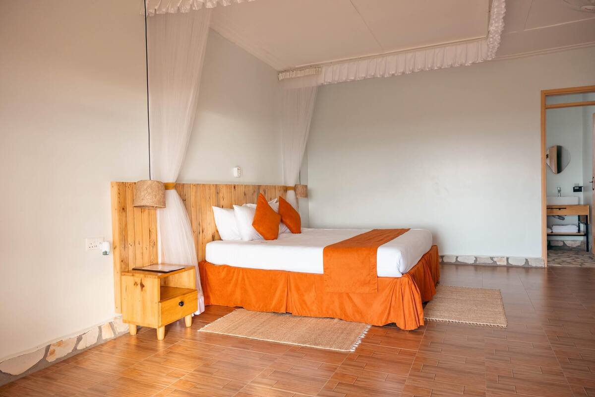 A bedroom with a wooden headboard, a bed with white and orange bedding, a small wooden nightstand, and a woven rug.