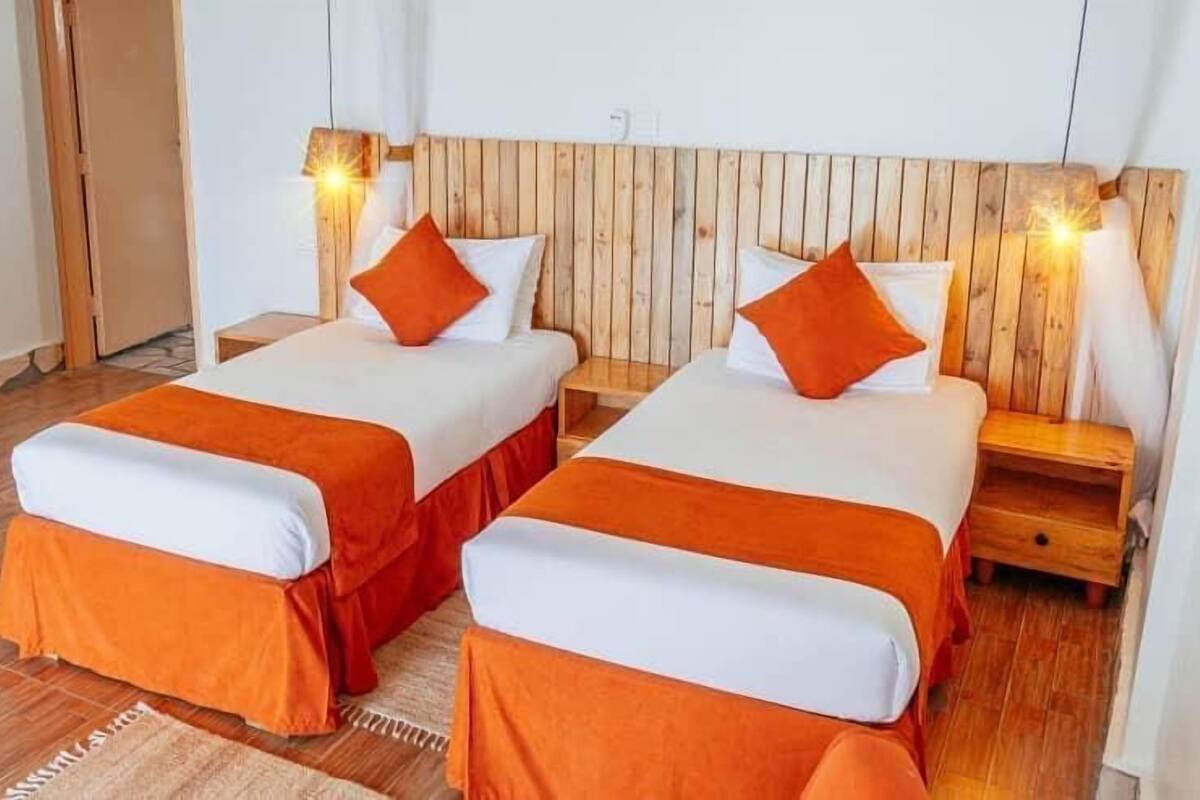 A room with two beds, each with an orange bedspread and white pillows. There are wooden headboards and nightstands with lamps.
