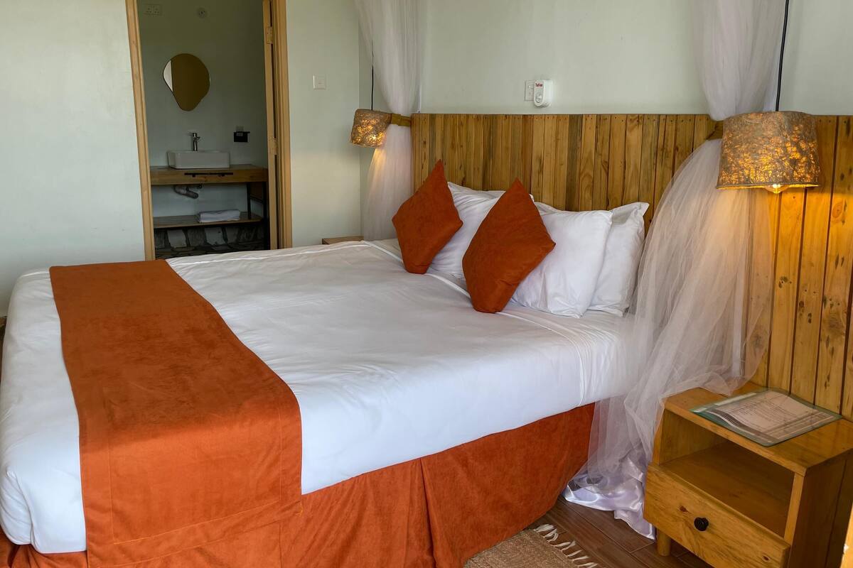 A neatly made bed with white and orange bedding, a wooden headboard, a bedside table with a lamp, and a mirror on the wall.