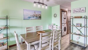 A dining area with a wooden table and white chairs, a painting of a lighthouse, a washing machine, and a shelf with decorative items.