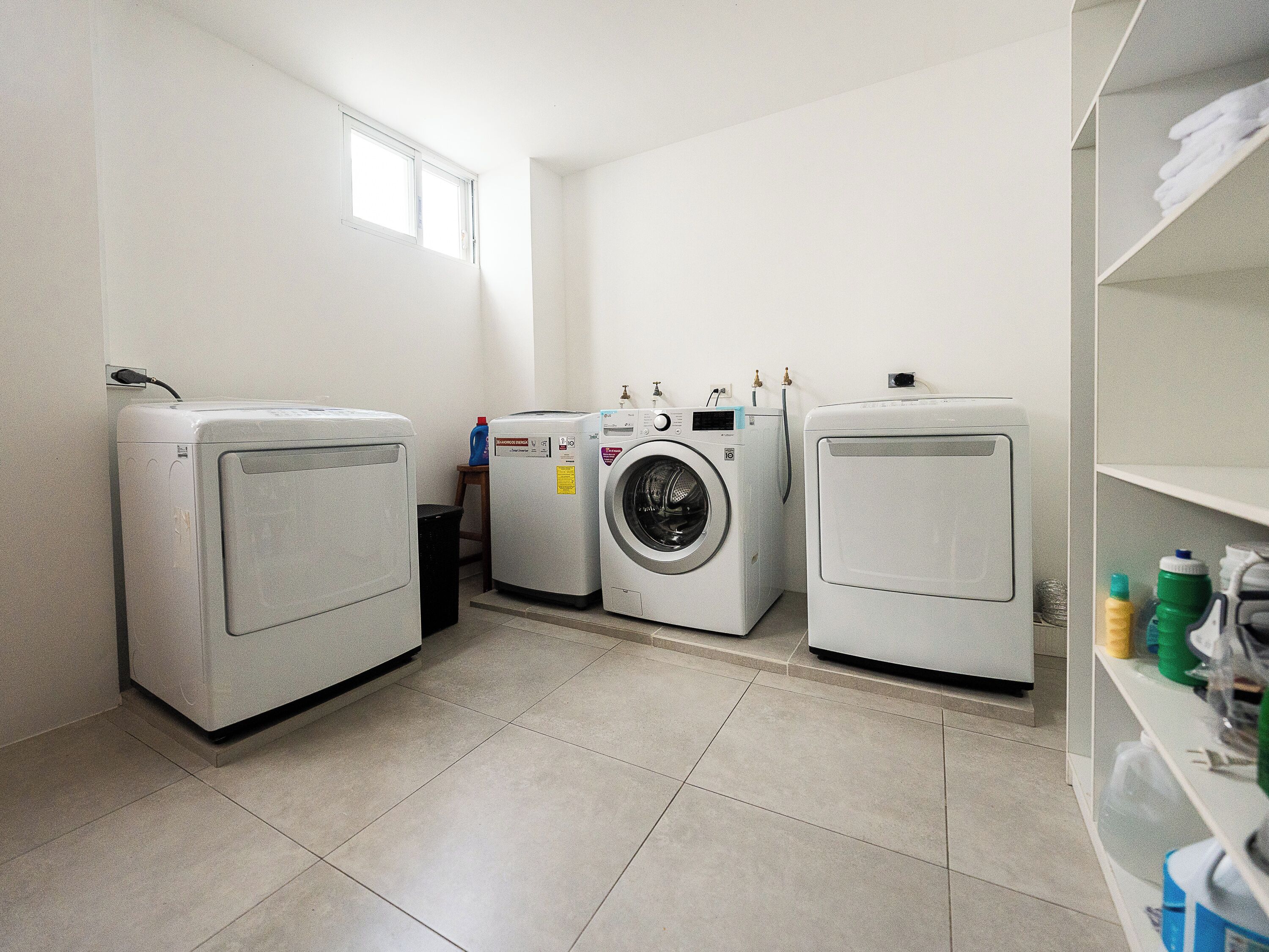 Una sala de bugaderia amb tres rentadora, un tamboret i una prestatgeria amb productes de neteja.