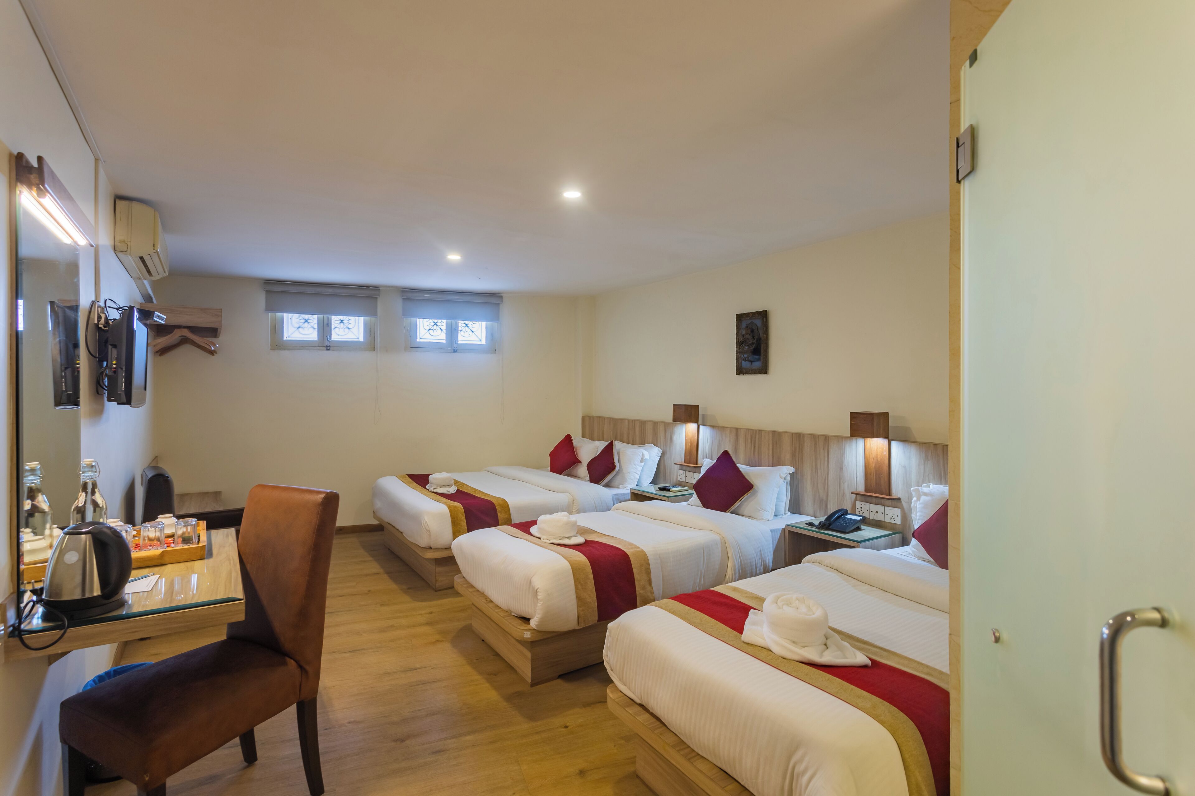 A hotel room with three beds, a desk with a chair, a minibar, and a window with blinds.