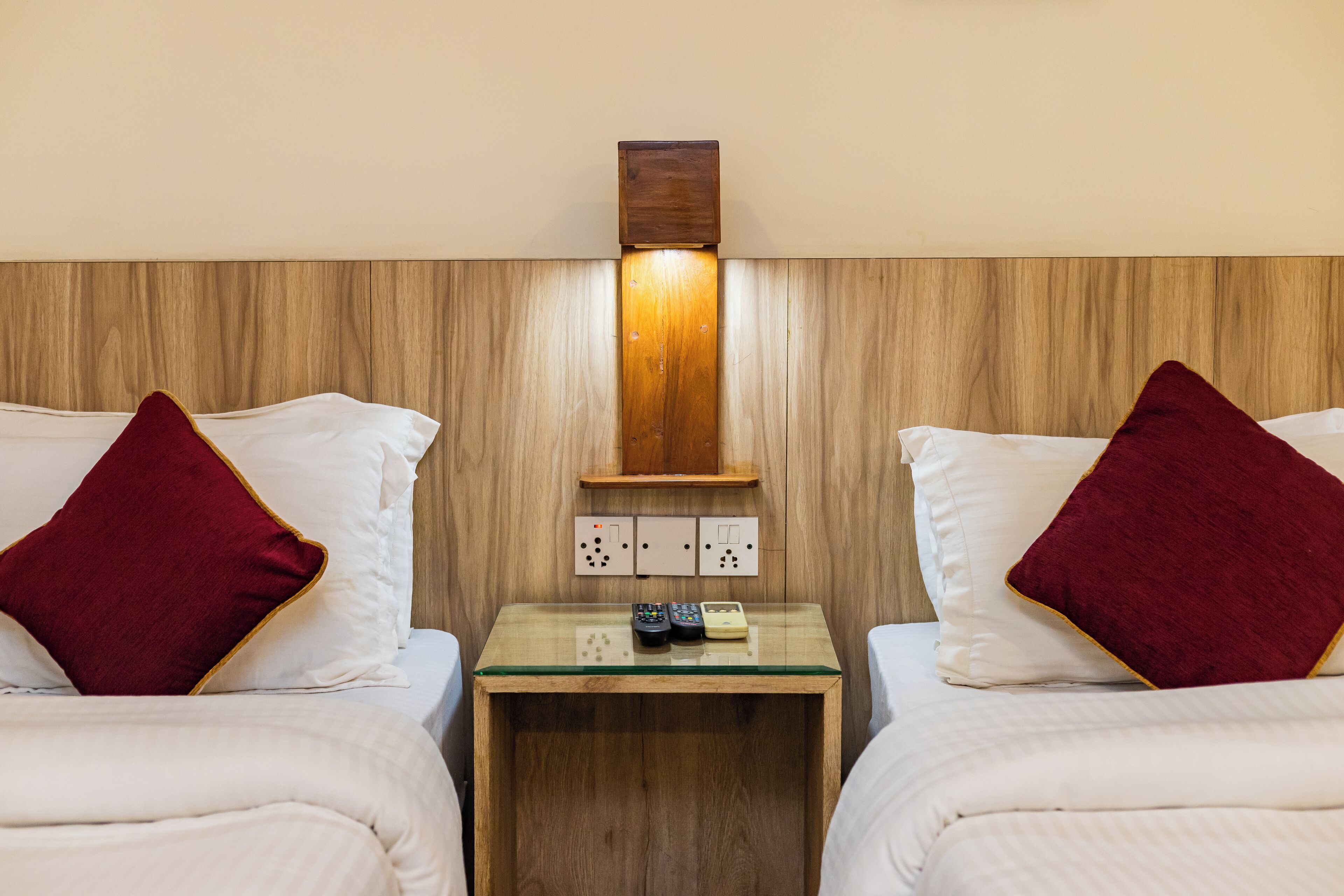 Habitació d'hotel amb dos llits, cada un amb roba de llit blanca i un coixí vermell decoratiu. Una petita tauleta de nit de fusta amb tapa de vidre conté un comandament a distància i un llibre.