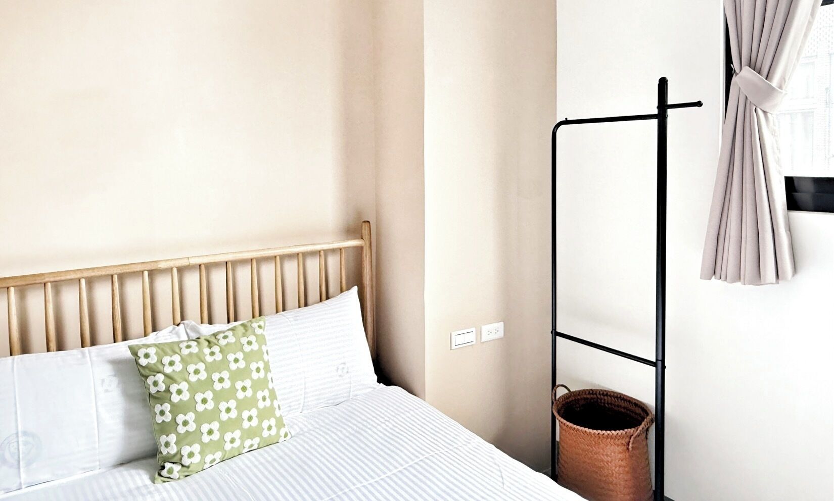 A bed with a wooden headboard, white bedding, and a green floral pillow. A black metal rack with a woven basket is against the wall.