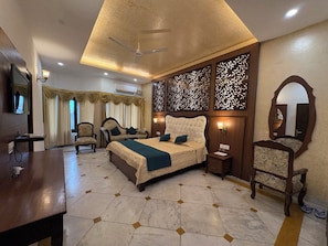 A hotel room with a bed, a desk, a chair, a TV, a ceiling fan, and decorative wall panels. - Yadavindra Gardens (Pinjore)