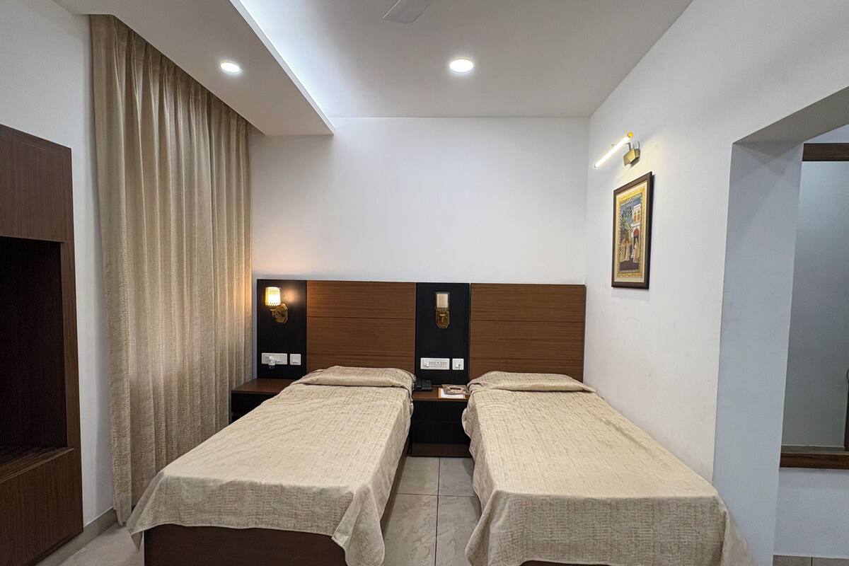 Quarto de hotel com duas camas de solteiro, cabeceira de madeira, mesa de cabeceira com abajur, quadro na parede e uma janela grande com cortinas.