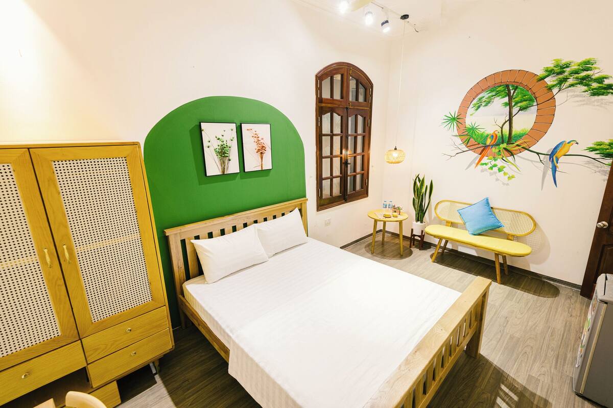 Een slaapkamer met een bed, kledingkast, groene wand, kunstwerk, raam, bankje en een kleine tafel met een plant.